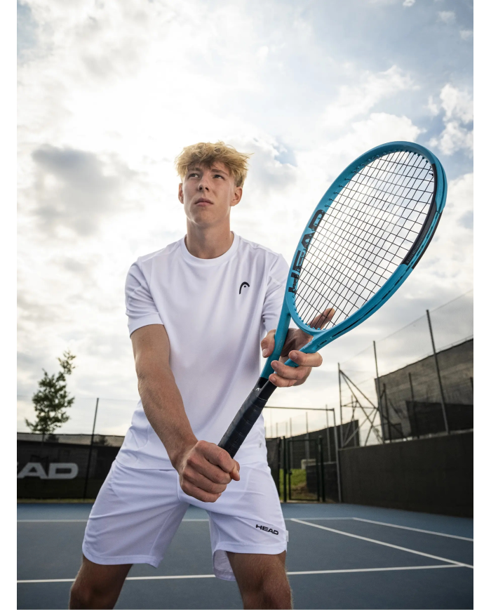 giovane uomo in bianco tiene racchetta da tennis azzurra in posizione pronta su campo all’aperto, cielo nuvoloso sullo sfondo, l